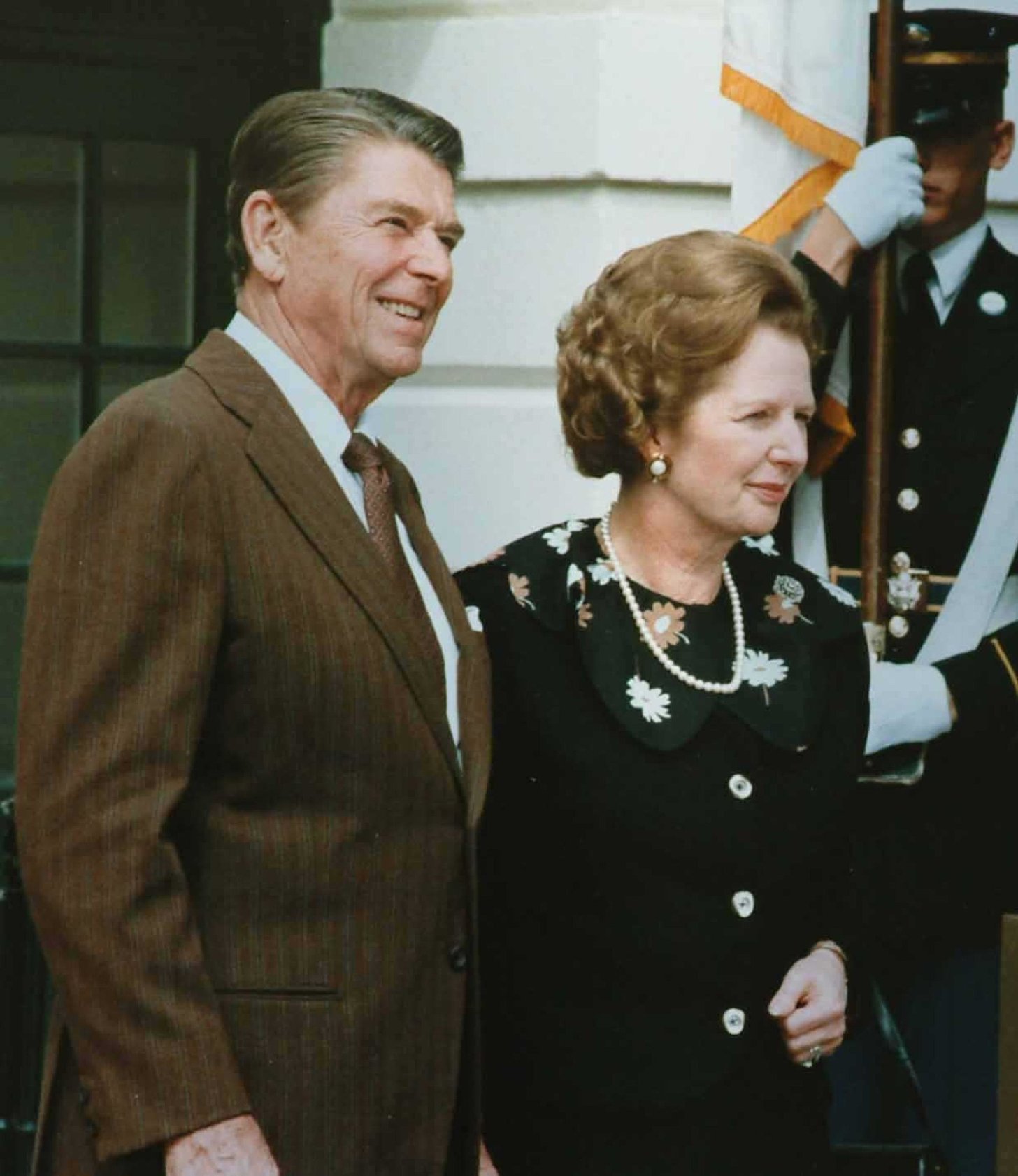 Prezident Ronald Reagan v hnědém obleku a Margaret Thatcherová v černých šatech s perlovým náhrdelníkem před Bílým domem, za nimi voják s vlajkou